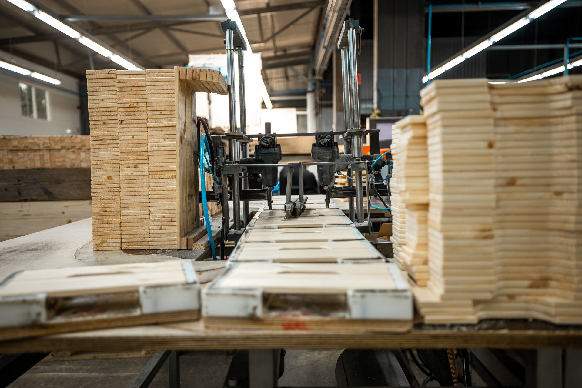 Industrial woodworking facility with stacks of wooden planks and pallets organized for processing, under bright industrial lighting, showcasing a busy and organized work environment